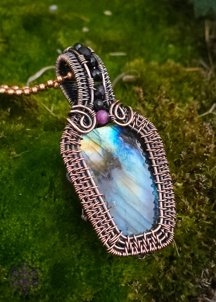 Copper wire wrapped labradorite coffin pendant with a red tourmaline accent stone at the top of the coffin to symbolize blood color, and then black spinel beads along the center of the bail. nice thick wrap style to create the ultimate statement piece. Labradorite has mainly blue flash with a few dark spots and yellow to orange colors. showing the left side.