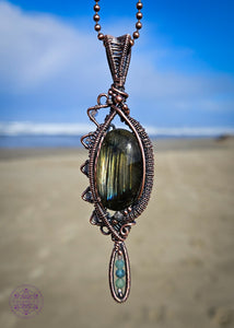 Copper wire wrapped necklace with an oval labradorite that has lines of yellow, green and blue flashes like the Norther Lights. On the left side there is a zig zag pattern with faceted rainbow moonstone bead with blue flash. The zig zag makes the pendant look like an eye when held sideways. At the base of the pendant is a long oval loop that encases small faceted smoky quartz and green tourmaline beads. Theres a copper coiling up the right side and copper accent beads throughout, truly a statement piece!