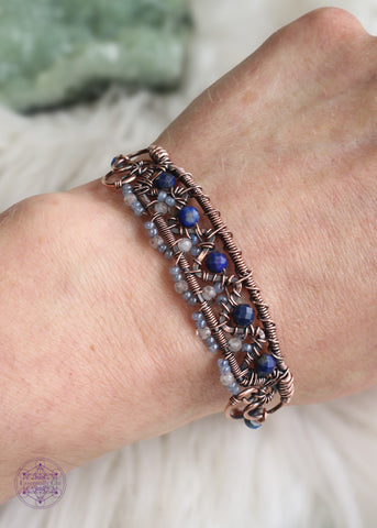 Copper wire wrapped bracelet features faceted lapis lazuli and moonstone woven between a zig zag design and added smoky blue seed beads for a filagree look. This bracelet is very detailed and sure to catch peoples eye! Shown worn on wrist with a white faux fur background. 