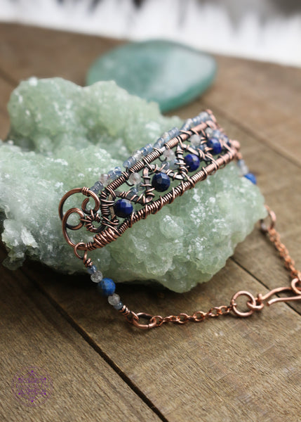 Copper wire wrapped bracelet features faceted lapis lazuli and moonstone woven between a zig zag design and added smoky blue seed beads for a filagree look. This view shows the  left side of the bracelet and the billow curls on the side as connector loops and the extention beaded detail of lapis lazuli, moonstone and seed bead combo. Bracelet is sitting on green crystal on a wooden table. 