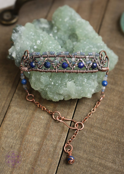 Copper wire wrapped bracelet features faceted lapis lazuli and moonstone woven between a zig zag design and added smoky blue seed beads for a filagree look. This view shows the front of the bracelet and the billow curls on the side as connector loops and the extention beaded detail of lapis lazuli, moonstone and seed bead combo. Bracelet is sitting on green crystal on a wooden table. 