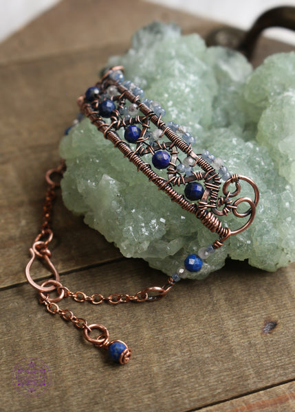 Copper wire wrapped bracelet features faceted lapis lazuli and moonstone woven between a zig zag design and added smoky blue seed beads for a filagree look. This view shows the right side of the bracelet and the billow curls on the side as connector loops and the extention beaded detail of lapis lazuli, moonstone and seed bead combo. Bracelet is sitting on green crystal on a wooden table. 
