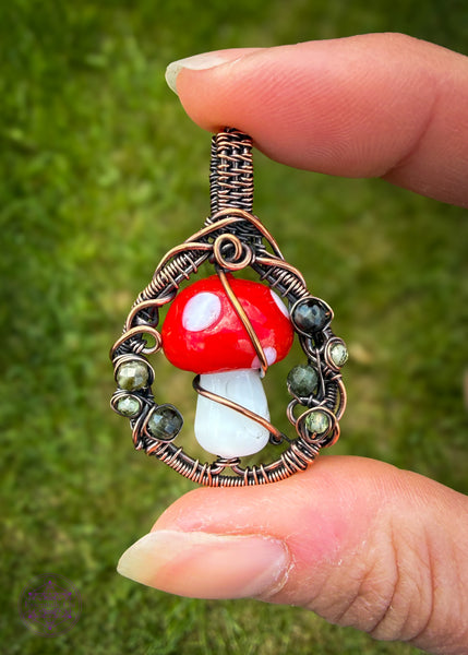 Copper wire wrapped design woven in a circle with a lampwork glass amanita mushroom in the center. The sides of the copper frame have faceted dark green tourmaline and peridot to simulate moss. Slight filagree slight fairy portal vibes. Held in hand with green grass in the background.