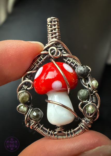 Copper wire wrapped design woven in a circle with a lampwork glass amanita mushroom in the center. The sides of the copper frame have faceted dark green tourmaline and peridot to simulate moss. Slight filagree slight fairy portal vibes. Held in hand with black leather in the background.