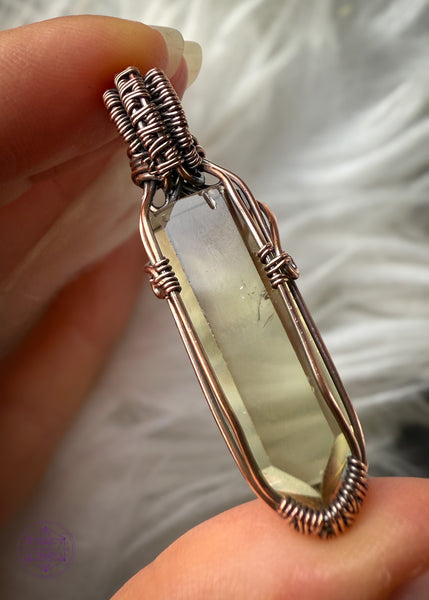 Copper wire wrapped Natural Citrine point pendant with a small smoky quartz bead in the bail. Held in fingers showing the back to show the natural inclusions of the wrap with a white faux fur background to show the details inside the crystal. There are two phantoms at the top of the crystal and the wrap doesn't cover any of the crystal to allow light to shine through the back. Some natural inclusions can be seen on the left middle side of the point.