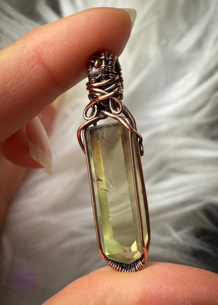 Copper wire wrapped Natural Citrine point pendant with a small smoky quartz bead in the bail. Held in fingers showing the right side of the wrap with a white faux fur background to show the details inside the crystal. There are two phantoms at the top of the crystal and the wrap doesn't cover any of the crystal to allow light to shine through the back. Some natural inclusions can be seen on the left middle side of the point.