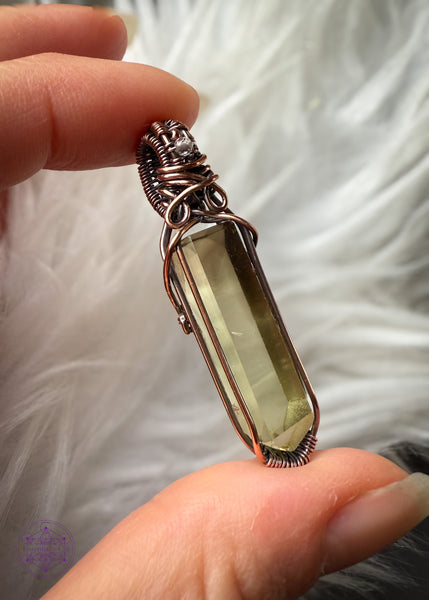 Copper wire wrapped Natural Citrine point pendant with a small smoky quartz bead in the bail. Held in fingers showing the left side of the wrap with a white faux fur background to show the details inside the crystal. There are two phantoms at the top of the crystal and the wrap doesn't cover any of the crystal to allow light to shine through the back. 