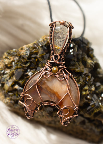 Copper wire wrapped Whale Tail or Mermaid Tail necklace with the fin in a natural agate with cascading colors of chocolate brown to caramel and butterscotch. The tail has swirls at the base to secure the stone in, and at the base of the bail is a natural colored tourmaline that matches the agate. The bail has a golden rutile upside-down tear drop and on top of that is a copper and two citrine beads. Shown on a white faux fur background leaning against an epidote cluster.