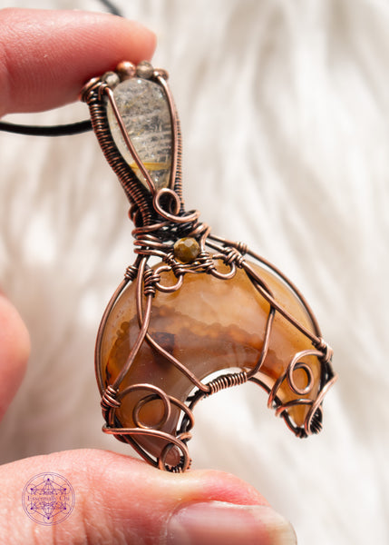 Copper wire wrapped Whale Tail or Mermaid Tail necklace with the fin in a natural agate with cascading colors of chocolate brown to caramel and butterscotch. The tail has swirls at the base to secure the stone in, and at the base of the bail is a natural colored tourmaline that matches the agate. The bail has a golden rutile upside-down tear drop and on top of that is a copper and two citrine beads. Shown on a white faux fur background held in hand showing the left angle.