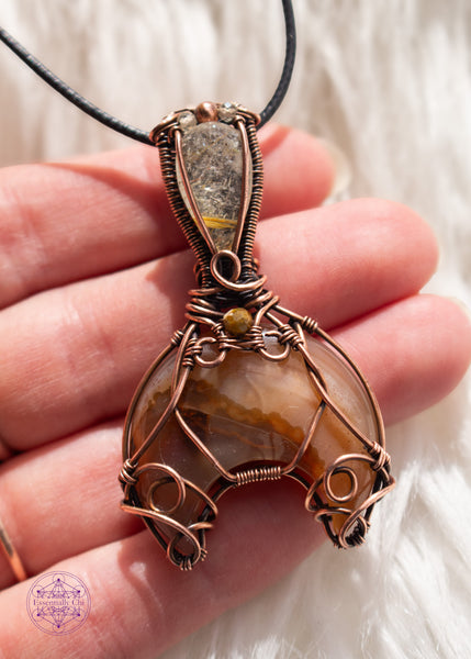 Copper wire wrapped Whale Tail or Mermaid Tail necklace with the fin in a natural agate with cascading colors of chocolate brown to caramel and butterscotch. The tail has swirls at the base to secure the stone in, and at the base of the bail is a natural colored tourmaline that matches the agate. The bail has a golden rutile upside-down tear drop and on top of that is a copper and two citrine beads. Shown on a white faux fur background held in hand for size reference.