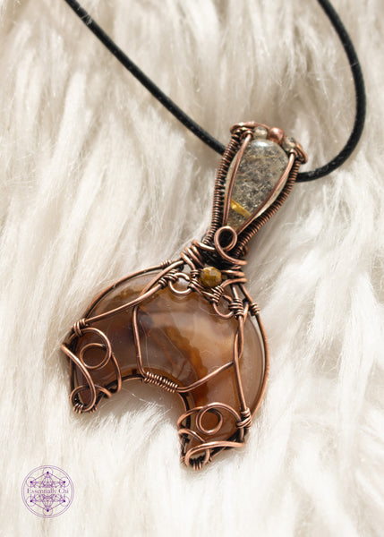 Copper wire wrapped Whale Tail or Mermaid Tail necklace with the fin in a natural agate with cascading colors of chocolate brown to caramel and butterscotch. The tail has swirls at the base to secure the stone in, and at the base of the bail is a natural colored tourmaline that matches the agate. The bail has a golden rutile upside-down tear drop and on top of that is a copper and two citrine beads. Shown on a white faux fur background.