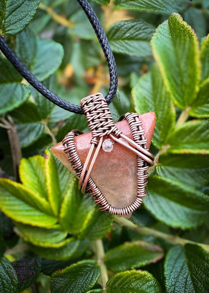 Small arrow shaped orange aventurine crystal pendant wrapped in copper with a woven design and a citrine crystal at the base of the bail. Shown over green leaves to help the orange in the pendant stand out. Hung on a black cotton cord.