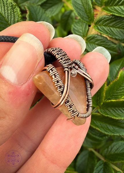 Small arrow shaped orange aventurine crystal pendant wrapped in copper with a woven design and a citrine crystal at the base of the bail. held showing the sides of the pendant and the durability of the wrap.