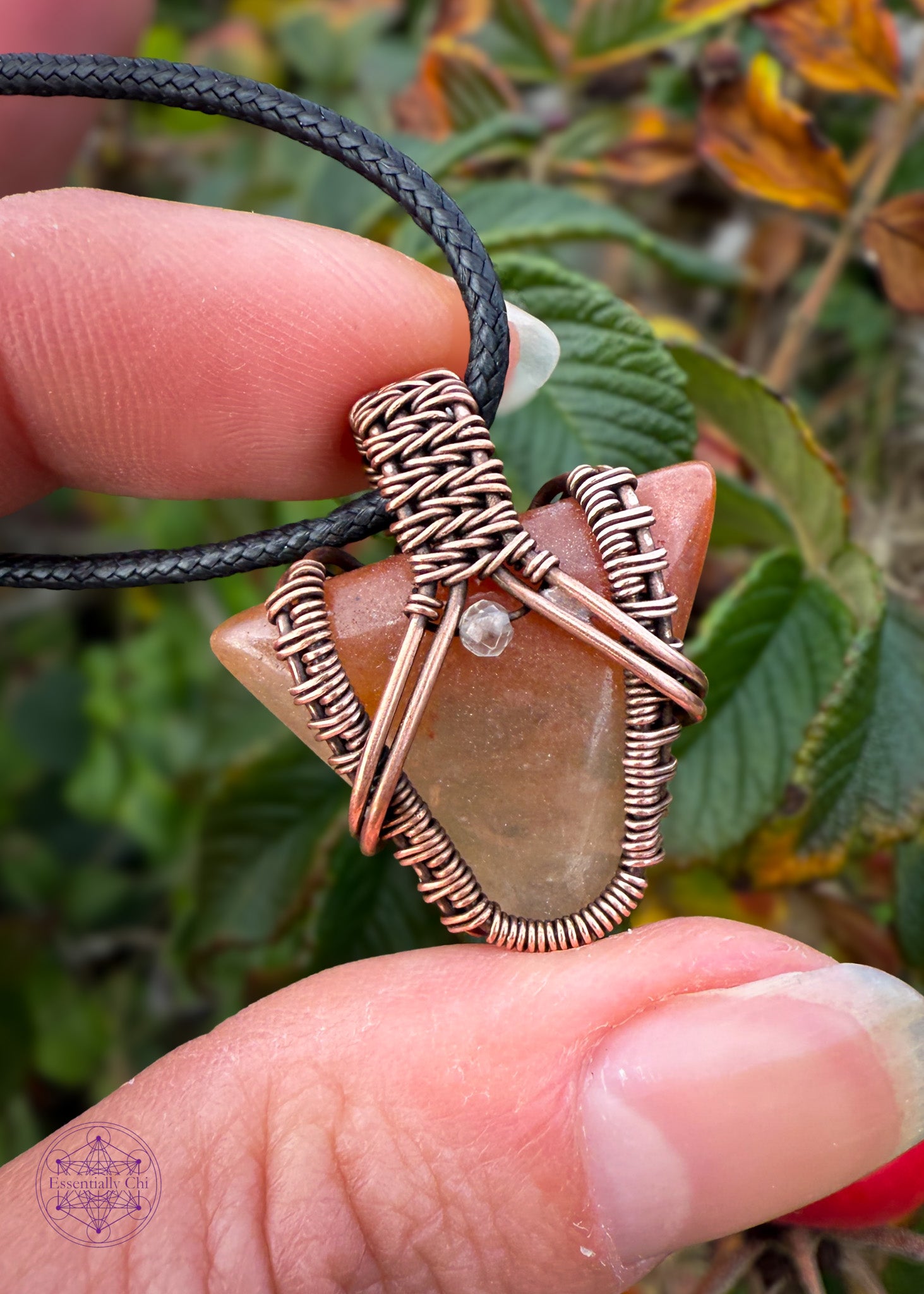 Small arrow shaped orange aventurine crystal pendant wrapped in copper with a woven design and a citrine crystal at the base of the bail. Held in fingers with autumn leaves of green and orange in the background.