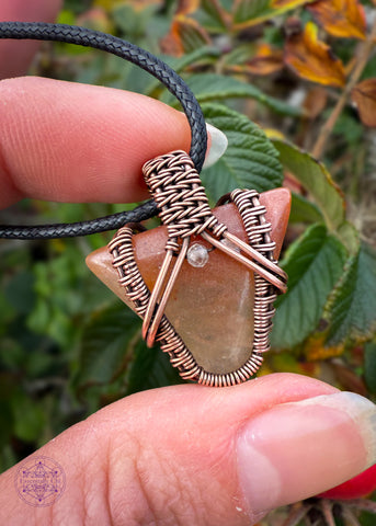 Small arrow shaped orange aventurine crystal pendant wrapped in copper with a woven design and a citrine crystal at the base of the bail. Held in fingers with autumn leaves of green and orange in the background.