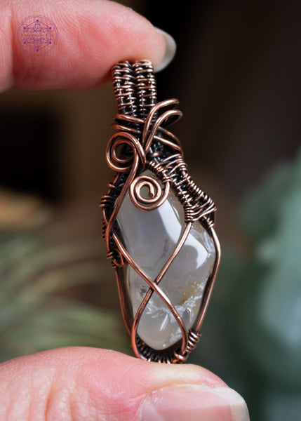 Oregon agate pendant with dendrite patterns and criss cross copper wire design. This is a long thin tumble stone with a few copper swirls at the base of the bail. Held in hand with a blurred background.