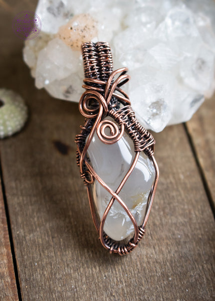 Oregon agate pendant with dendrite patterns and criss cross copper wire design. This is a long thin tumble stone with a few copper swirls at the base of the bail. Shown sitting on a wooden table leaning against an apophyllite crystal.