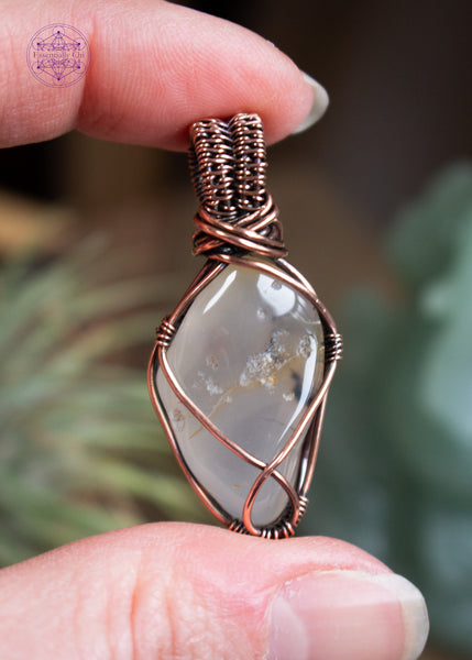 Copper-wrapped crystal pendant with an Oregon agate tumble stone with a simple copper wire wrap. held between fingers with a blurred natural background.