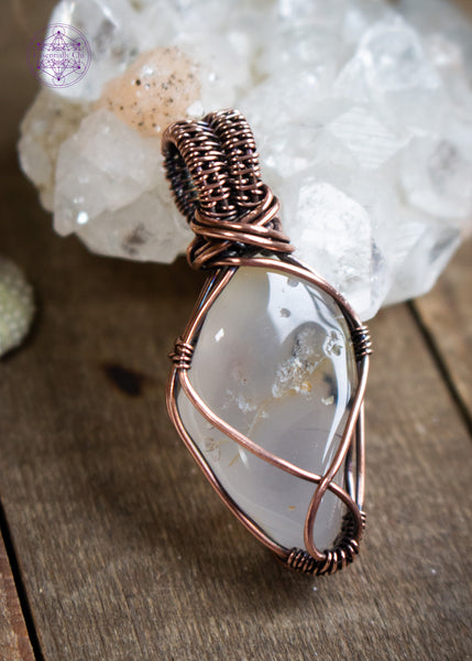Copper-wrapped crystal pendant with a smooth  Oregon agate tumble stone with a simple copper wire wrap. Sitting on a wooden table leaning against an apophyllite crystal.