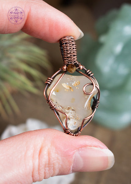 Triangle shapped Oregon agate pendant with copper wire wrap and brown tourmaline accent crystal at the base of the bail. Held in hand with crystals and airplants in the blurred background.