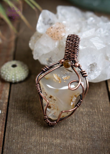 Triangle shapped Oregon agate pendant with copper wire wrap and brown tourmaline accent crystal at the base of the bail. Shown leaning on an apophyllite crystal on a wooden table with a small urchin shell next to it.