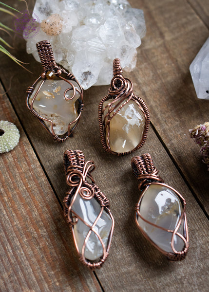 Oregon agate copper wire wrapped pendants from Yachats Oregon, four different styles shown sitting on a wooden board with crystals and a tiny sea urchin shell around it.