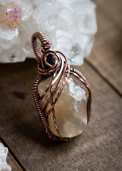 Oregon agate small nugget pendant wrapped in copper wire with a rosette detail at the base of the bail and three swoops going down the left side. Has smooth but bumpy texture perfect for those who like texture in their jewelry to play with. Shown sitting on a wooden board with an apophyllite crystal in the background.