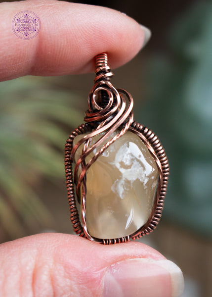 Oregon agate small nugget pendant wrapped in copper wire with a rosette detail at the base of the bail and three swoops going down the left side. Has smooth but bumpy texture perfect for those who like texture in their jewelry to play with. Held in hand with crystals and airplants blurred in the background.