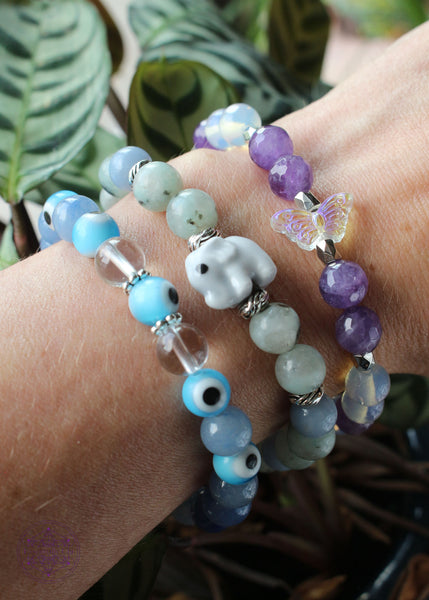 Three bracelets shown on wrist. One light blue evil eye with blue aventurine, a kiwi jasper and angelite with a lampwork elephant focal bead, and a faceted amethyst with opalite and an acrylic iridescent butterfly. 