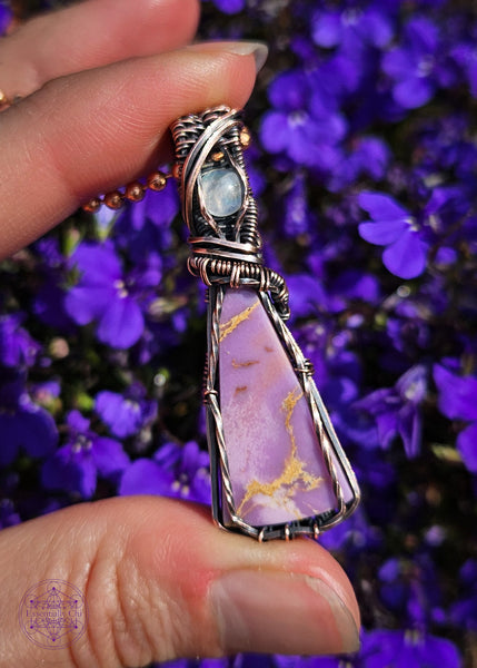 Copper wire wrapped Phosphosiderite crystal with a flashy moonstone accent crystal in the bail. The Phosphosiderite is a lavender colored crystal with some beige inclusions throughout in a thin tall triangle shape, wrapped to show off as much as the crystal as possible and in an elegant design.