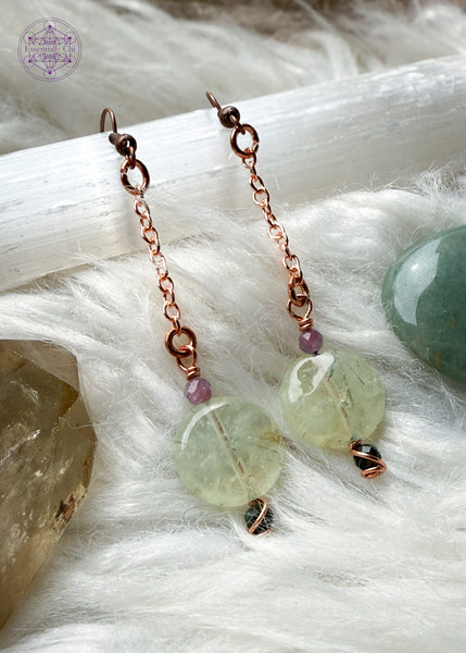 Gorgeous glowing prehnite dangle earrings hang from a long copper chain with green and pink tourmaline faceted beads and copper wire swirled around them. Hung on brass ear hooks gives them a total length of 3". Shown on selenite and white faux fur and crystals.