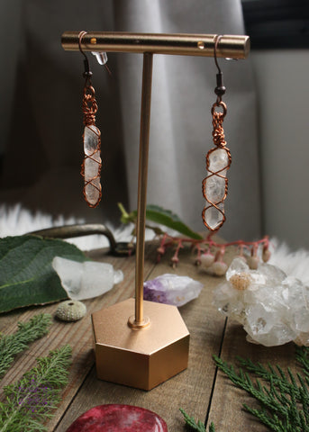 Copper wire earrings in a twisted cage style, enclosed around thin raw quartz points. Hanging from an earring display with the light shining through behind them. Foliage and crystals were displayed on the wooden table.