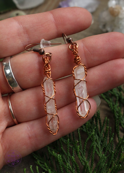Copper wire earrings in a twisted cage style enclosed around thin raw quartz points. Sitting on a wooden table with crystals and foliage around the background.