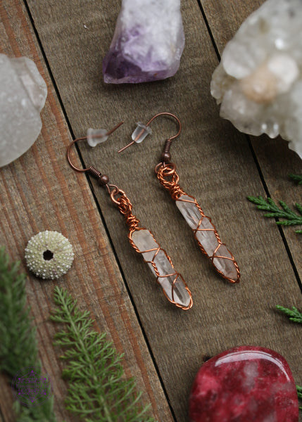 Copper wire earrings in a twisted cage style enclosed around thin raw quartz points. Sitting on a wooden table with crystals and foliage around the background.