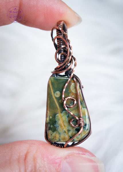 An organically shaped rainforest rhyolite stone wrapped in copper wire with a flowing wire swirl cascading down the right side, designed to highlight the natural orb patterns within the rhyolite. Held in hand with a white faux fur background,
