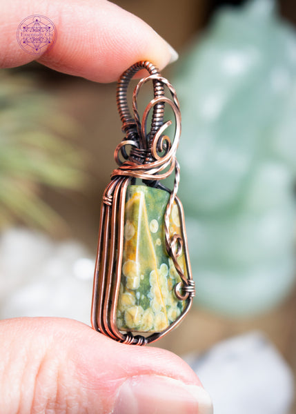 An organically shaped rainforest rhyolite stone wrapped in copper wire with a flowing wire swirl cascading down the right side, designed to highlight the natural orb patterns within the rhyolite. Shwoing the left side details of 4 straight square shaped wire to hug the side of the crystal.
