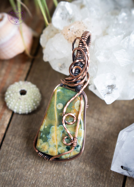 An organically shaped rainforest rhyolite stone wrapped in copper wire with a flowing wire swirl cascading down the right side, designed to highlight the natural orb patterns within the rhyolite. Shown laying on a wooden table with crystals and shells around it.