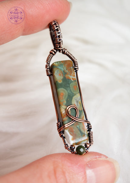 Copper wire wrapped rainforest rhyolite longer rectangular stone with a simple bail and a loop design at the base, accented with small copper beads and a faceted green tourmaline bead. Shown held in hand with a white faux fur background.