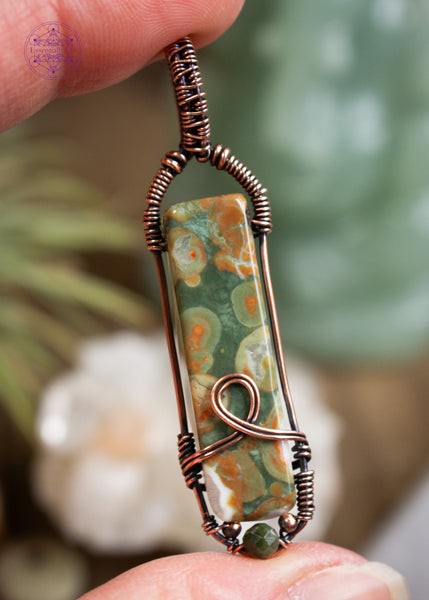 Copper wire wrapped rainforest rhyolite longer rectangular stone with a simple bail and a loop design at the base, accented with small copper beads and a faceted green tourmaline bead. Shown held in hand with a blurred background of crystals.