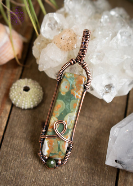 Copper wire wrapped rainforest rhyolite longer rectangular stone with a simple bail and a loop design at the base, accented with small copper beads and a faceted green tourmaline bead. Shown sitting on a wooden table leaning against an apophyllite crystal and a urchin shell next to it.