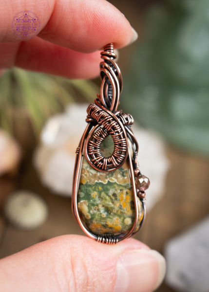 my favorite design features a copper wire wrapped rainforest rhyolite with a woven loop at the base of the bail, soft wire swoops along the front of the bail. A natural indent on the right of the stone holds a brown tourmaline accent with copper beads, adding depth and detail. Held in hand with a blurred background of crystals on a table.