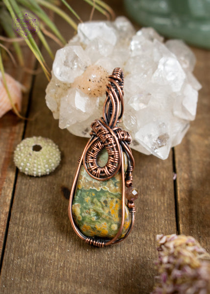 my favorite design featuring a copper wire wrapped rainforest rhyolite with a woven loop at the base of the bail, soft wire swoops along the front of the bail. A natural indent on the right of the stone holds a brown tourmaline accent with copper beads, adding depth and detail. Shown sitting on a wooden table leaning against an apophyllite crystal.