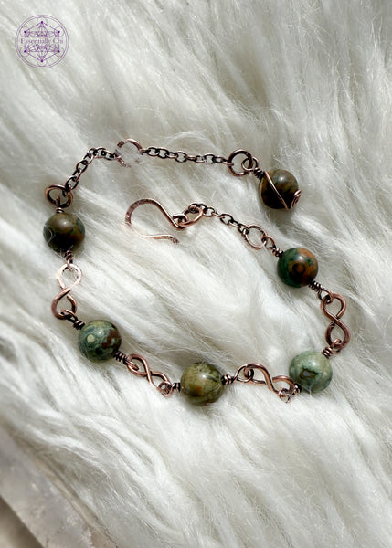 Copper wire wrapped bracelet features 8mm rainforest rhyolite beads individually wrapped with figure 8 Infinity links in-between for an elegant bracelet. Showing the whole bracelet on a faux white fur blanket to see hook clasp and beaded extension chain and rhyolite charm.