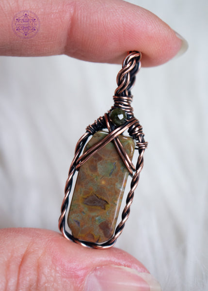 This dainty copper wire wrapped rainforest  rhyolite has a small rectangular stone framed with a braided border and a criss-cross detail at the top. Finished with a faceted green tourmaline bead at the base of the bail. Held in hand with a white faux fur background.