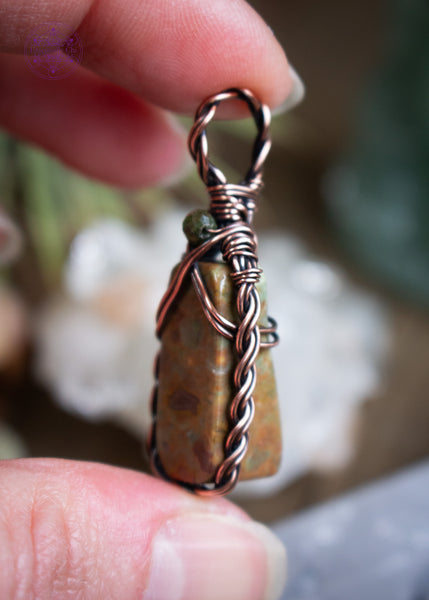 This dainty copper wire wrapped rainforest  rhyolite has a small rectangular stone framed with a braided border and a criss-cross detail at the top. Finished with a faceted green tourmaline bead at the base of the bail. Held in hand showing the side details with a background blurred.