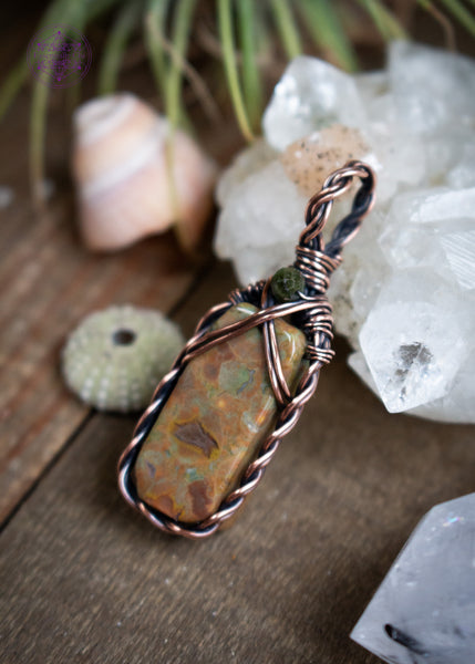 This dainty copper wire wrapped rainforest  rhyolite has a small rectangular stone framed with a braided border and a criss-cross detail at the top. Finished with a faceted green tourmaline bead at the base of the bail. Shown sitting on a wooden table with crystals and shells next to it.