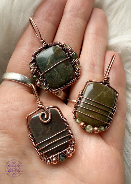 Three dainty rainforest rhyolite (jasper) pendants in hand to show the difference in the three. One is dark green with hints of red with beads in tourmaline, copper and peridot. The second is an light olive green with beads along the bottom of light green tourmaline and peridot. The third pendant is a dark green with maroon spots and a copper swirl at base of bail and beads of copper and a dark green faceted tourmaline. 