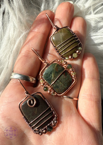 Three dainty rainforest rhyolite (jasper) pendants in hand to show the difference in the three. One is dark green with hints of red with beads in tourmaline, copper and peridot. The second penant is a dark green with maroon spots and a copper swirl at base of bail and beads of copper and a dark green faceted tourmaline. The third pendant is a light olive green with beads along the bottom of light green tourmaline and peridot. 
