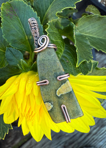 This rainforest rhyolite pendant is wrapped in copper wire with a simple prong design, one prong at the base, and one on each side. The bail is a simple narrow bail with a delicate swirl at the base to hold the pendant together. Simple elegant olive green stone with map like creamy yellow spots within the stone. Shown on a yellow dahlia to show off the gorgeous green color in the pendant. 