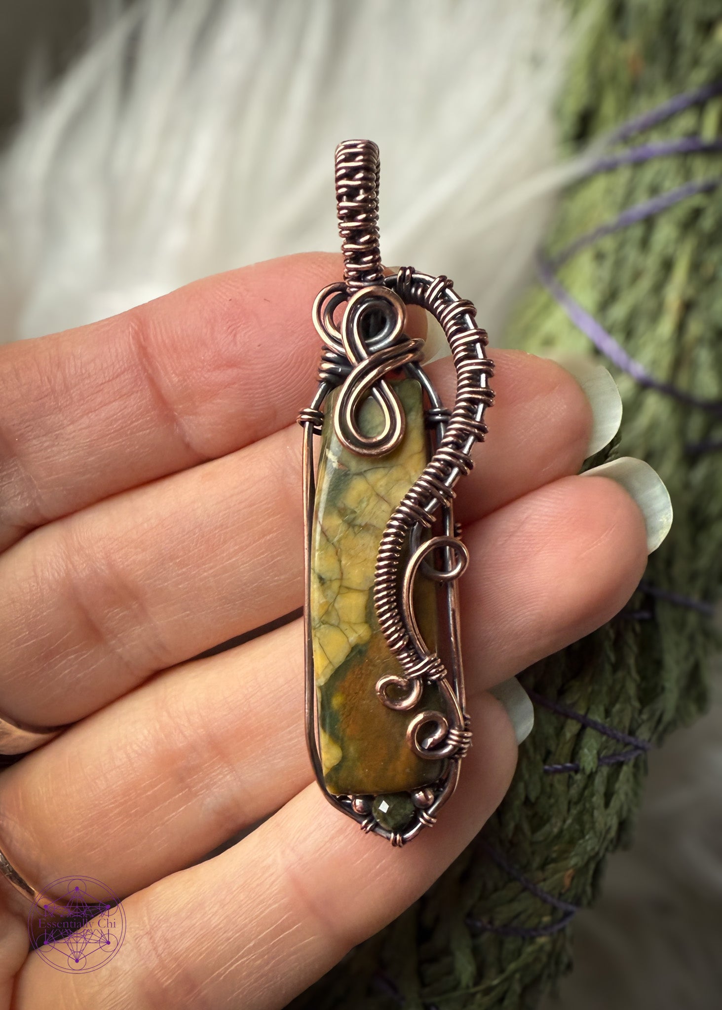 Rainforest Rhyolite Pendant wrapped in copper wire. This pendant is a long thin jasper (rhyolite) with dark mossy green and olive yellow/green spots along the top of the crystal and dark brown near the bottom. The copper curls around the face of the stone to show the details of the green and brown, and adding curls at the bottom with the wire like fern curls. The base of the pendant I added the Green tourmaline bead to fill in the point I added at the bottom with wire. The bail is simple narrow weave. 